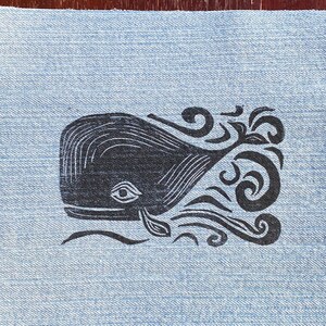 May include: A black and white whale illustration printed on a blue denim fabric patch. The whale is facing left and has a stylized design with swirls and waves.