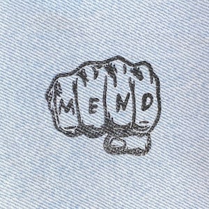 May include: A square, light blue denim patch with a black illustration of a clenched fist. The word "MEND" is printed across the knuckles in black capital letters. The patch is on a dark brown wooden surface.
