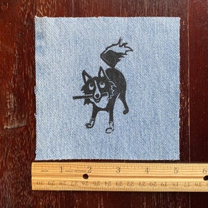 Op de afbeelding: Een zwart-witte illustratie van een hond met een stok in zijn bek, gedrukt op een blauwe denim stoffen patch.