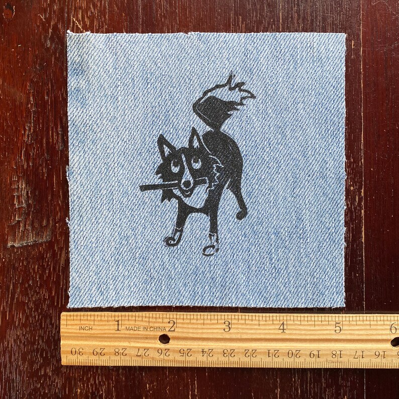 Op de afbeelding: Een zwart-witte illustratie van een hond met een stok in zijn bek, gedrukt op een blauwe denim stoffen patch.