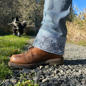 Puede incluir: Un par de pantalones vaqueros azules con un diseño de olas bordado en el dobladillo. Los vaqueros se llevan con botas de cuero marrón.