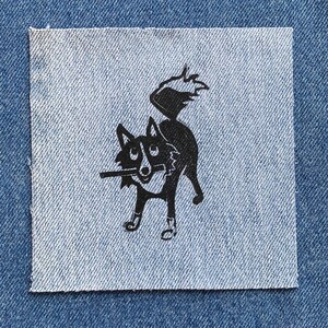 Op de afbeelding: Een zwart-witte illustratie van een hond met zijn staart in de lucht, gedrukt op een blauwe denim patch.