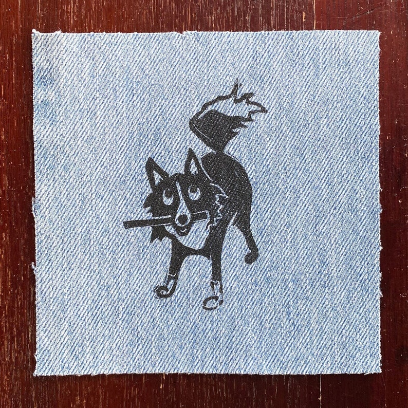 Op de afbeelding: Een zwart-witte illustratie van een hond met zijn tong naar buiten, gedrukt op een blauwe denim patch.