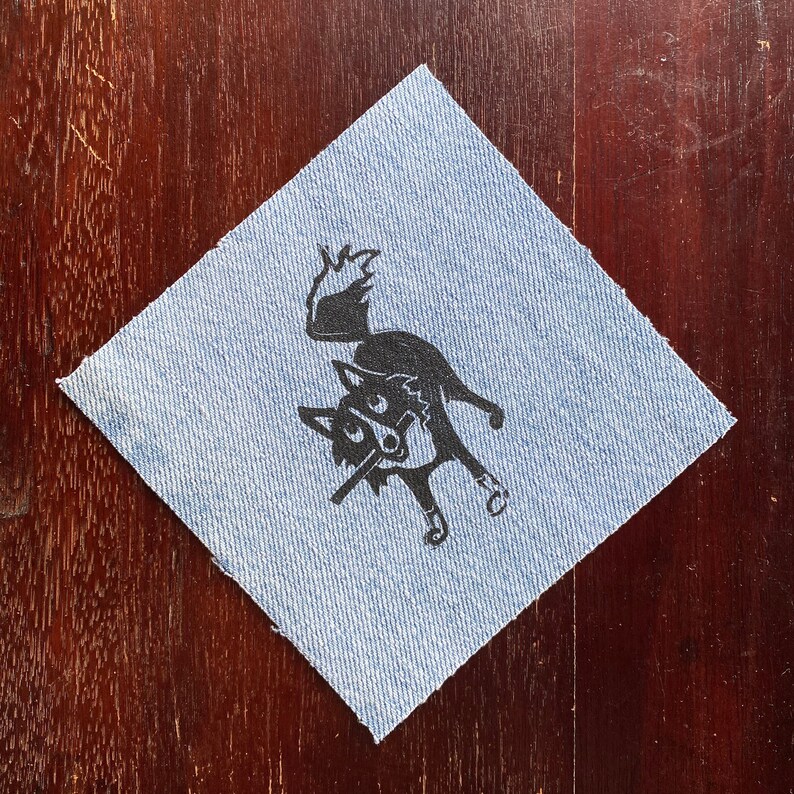 Op de afbeelding: Een zwart-witte illustratie van een wolf of vos met een lange snuit en grote oren, gedrukt op een blauwe denim patch.