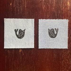 Puede incluir: Dos cuadrados pequeños de tela vaquera azul con un diseño de pájaro negro impreso en cada uno. El pájaro está estilizado y tiene un ala extendida.