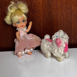 Könnte beinhalten: Eine kleine Puppe mit blondem Haar in zwei Knoten, bekleidet mit einem rosa Kleid mit weißer Spitzenborte, neben einem kleinen, weißen, lockigen Plüschlamm mit rosa Ohren und rosa Nase, beide auf einer weißen Oberfläche stehend.