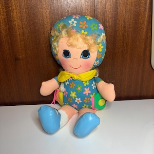 以下が含まれることがあります： 青い花柄のドレスと帽子を着たヴィンテージドール。人形はブロンドの巻き毛、バラ色の頬、青い目をしています。黄色いリボンと青い靴を履いています。人形は足を伸ばして座っています。