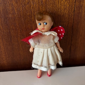 May include: A vintage doll with brown hair, blue eyes, and red lips. The doll wears a cream-coloured dress with a sheer overlay and red polka-dot wings. The doll's shoes are red.
