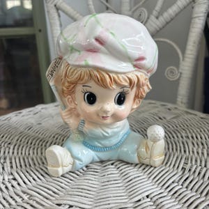 May include: A ceramic figurine of a child wearing a pink and green hat and blue overalls holding a golf club and a golf ball. The figurine is a planter.