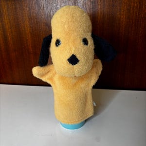 May include: A yellow plush dog hand puppet with black ears and a black nose. The puppet has a light blue base and is designed for children's play. The puppet's eyes are black and shiny.