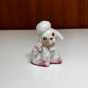 Puede incluir: Pequeña figura de caniche de cerámica blanca, con detalles rosas en las patas y orejas. El caniche tiene ojos negros, nariz rosa y un pequeño lazo. La figura está sentada y tiene una textura rizada.