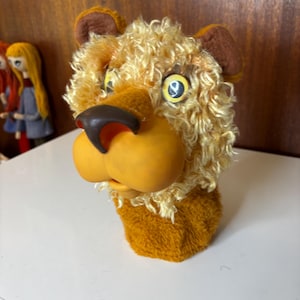 May include: A plush lion head puppet with a tan and brown color scheme. The lion has a large orange nose, yellow eyes, and a shaggy mane. The puppet is made of soft, textured materials and is designed to be worn on the hand.