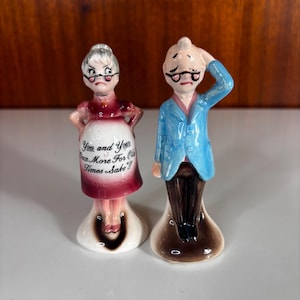 May include: Two ceramic figurines depicting an elderly couple. The figure on the left wears a red dress with an apron featuring the text "You and Your Once More For Old Times Sake!!". The figure on the right wears a blue jacket and brown trousers.