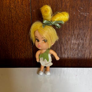 Könnte beinhalten: Eine Vintage-Puppe mit gelbem Haar, das zu einem Dutt und Pony frisiert ist, und einer grünen Schleife. Die Puppe trägt ein grünes und weißes Kleid und silberne Schuhe. Die Puppe hat große Augen und einen Schmollmund. Die Puppe ist ein Sammlerstück.