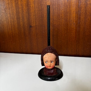 May include: A vintage doll head on a black stand. The doll head has painted features, including blue eyes and red lips. The doll is wearing a maroon hood and collar. The stand is round and black, with a tall black rod extending upwards.