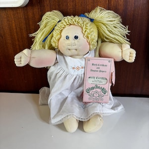 May include: A vintage Cabbage Patch Kids doll with blonde yarn pigtails tied with blue ribbons. The doll wears a white dress with floral embroidery and lace trim. It holds a pink birth certificate that reads "Elana Sissy."