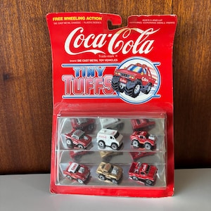 Puede incluir: Un juego de coches de juguete Coca-Cola Tiny Tuffs. El embalaje rojo y blanco presenta el logotipo de Coca-Cola y las palabras "Tiny Tuffs". El juego incluye varios vehículos de juguete de metal fundido a presión.