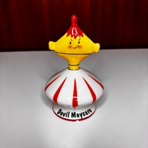 May include: A ceramic figurine featuring a yellow head with a red pointed top, a smiling face, and a white base with red vertical stripes. The base displays the black text Devil Maycare. The object has a glossy finish and a rounded, symmetrical shape.