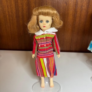 Może przedstawiać: Zabytkowa lalka z blond włosami i niebieskimi oczami, ubrana w czerwoną, zieloną i ż&oacute;łtą kurtkę i sp&oacute;dnicę w paski. Kurtka ma biały kołnierz i zielone guziki. Lalka stoi na przezroczystym plastikowym stojaku.