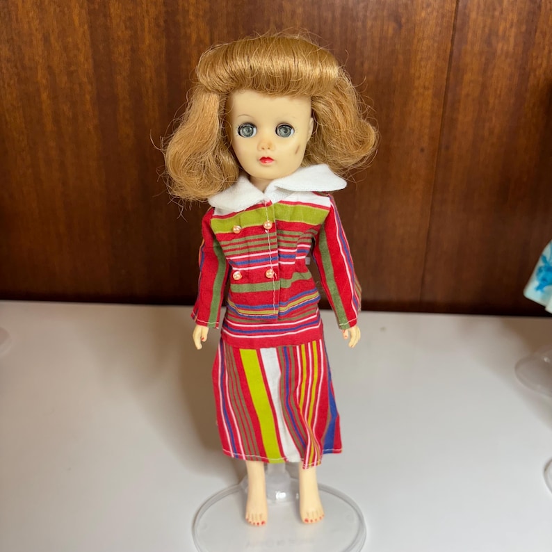 Może przedstawiać: Zabytkowa lalka z blond włosami i niebieskimi oczami, ubrana w czerwoną, zieloną i ż&oacute;łtą kurtkę i sp&oacute;dnicę w paski. Kurtka ma biały kołnierz i zielone guziki. Lalka stoi na przezroczystym plastikowym stojaku.
