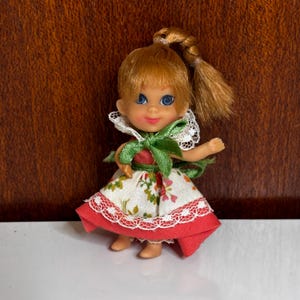 May include: A small doll with light brown hair styled in a high ponytail, blue eyes, and a smiling expression. The doll wears a white dress featuring a floral pattern, a green ribbon tied at the neck, a white lace collar, and a solid red hem.