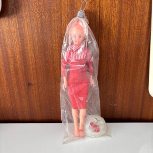 May include: A vintage doll in a clear plastic bag. The doll is wearing a red outfit with a matching scarf and shoes. The doll has blonde hair and blue eyes. A small, white, circular stand with red accessories is at the bottom of the bag.