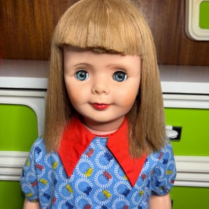 Puede incluir: Una muñeca vintage con cabello rubio y ojos azules. La muñeca viste un vestido azul con cuello rojo y un estampado de trenes. La cara de la muñeca tiene una expresión pintada con labios rojos y mejillas rosadas. Un juguete clásico.