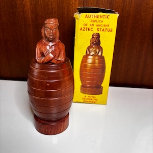 以下が含まれることがあります： 腕を組んだ人物の茶色の木製フィギュアで、樽型の台座から出ています。その横には、AUTHENTIC REPLICA OF AN ANCIENT AZTEC STATUE A NOVEL DECORATIVE ORNAMENTという文字が書かれた黄色い段ボール箱があります。