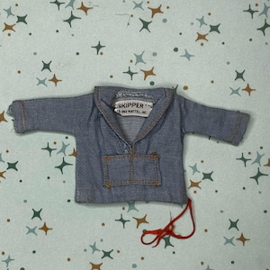 May include: A light blue denim-style doll jacket with long sleeves, a collar, and a front pocket. The interior label reads "SKIPPER T.M. 1963 MATTEL, INC." A loose red thread hangs from the bottom right corner of the garment, which features visible brown stitching.