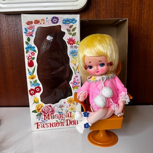 May include: A blonde doll with large blue eyes wearing a pink long-sleeved dress with white lace trim and two white pom-poms, seated in an orange pedestal chair. Beside it is a white box with floral illustrations and the text Musical Fashion Doll.