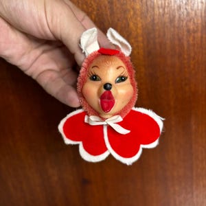 May include: A vintage-style ornament featuring a stylized animal face with a red fuzzy hood, white ears, and a red tongue. The figure wears a red velvet cape with white scalloped trim and a small white ribbon bow tied at the center neck area.