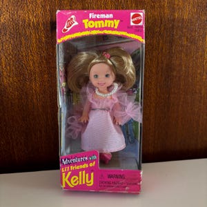 May include: A Fireman Tommy doll in its original packaging. The doll has blonde hair, blue eyes, and is wearing a pink dress with a pearl necklace. The packaging is pink and yellow with the Mattel logo.