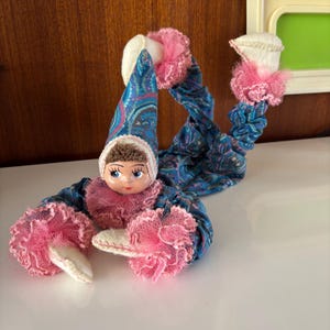 Könnte beinhalten: Eine Vintage-Puppe mit bemaltem Gesicht, gekleidet in einem blau-paisleymusternden Outfit mit rosa Besatz. Die Puppe hat einen langen, spitzen Hut und passende Beine. Die Arme und Beine der Puppe sind beweglich.