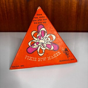 Puede incluir: Una caja triangular naranja para un Pixie Bow Maker. El frente presenta un gráfico de un lazo de cinta blanca sobre una forma rosa, con el texto: Have fun, make bow after bow the easy way, PIXIE BOW MAKER, MAG-NIF, INC. y PAINESVILLE, OHIO.