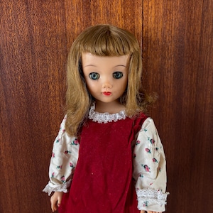 May include: A vintage doll with blonde hair and blue eyes. The doll wears a red velvet dress with white sleeves adorned with a floral pattern. The dress has a white lace collar. The doll is a collectible item.