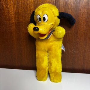 Könnte beinhalten: Ein Vintage-Pluto-Plüschtier. Das Spielzeug hat einen gelben Kopf und Körper mit schwarzen Ohren und einer schwarzen Nase. Pluto hat große, weiße Augen und einen roten Mund. Ein farbenfroher gestreifter Kragen ist um seinen Hals. Das Plüschtier steht aufrecht.