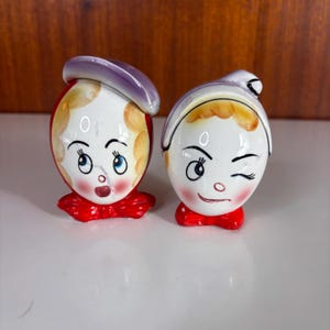 May include: A pair of ceramic salt and pepper shakers shaped like cartoon heads. One shaker features a purple hat, blonde hair, and a surprised expression. The other shaker has a white hat, blonde hair, and a winking expression with a red bow.