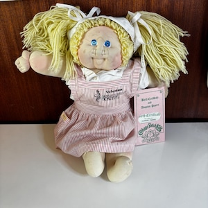May include: A Cabbage Patch Kids doll with blonde yarn hair in pigtails, blue eyes, and a smiling face. The doll wears a pink and white striped dress with a white apron and the words "Volunteer Park-Central Hospital" printed on it. A birth certificate is included.