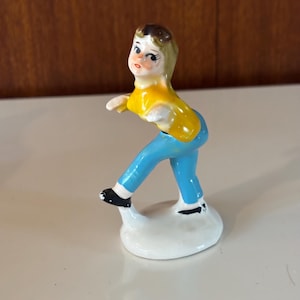 May include: A vintage ceramic figurine of a person in a skating pose. The figurine is wearing a yellow shirt, blue pants, and black shoes. The figure is standing on a white base. The figurine has brown hair and rosy cheeks.