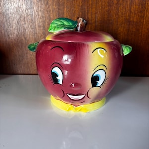 May include: A ceramic apple-shaped jar with a lid, featuring a glossy red and yellow finish. The jar has a cartoon-style face with large blue eyes, long eyelashes, and a smiling mouth. A green leaf is attached to the brown stem on the lid.