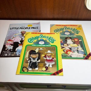 May include: Three vintage books featuring Cabbage Patch Kids and Little People Pals. The books have colorful covers with illustrations of dolls and outfits. The titles include "Pony & Western Wear" and "Designer Clothes."