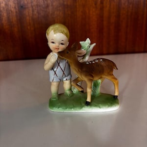 May include: A ceramic figurine featuring a small child with blonde hair wearing a white outfit with a brown diamond pattern, standing next to a brown fawn with white spots on its back. Both figures are positioned on a green base with a plant.