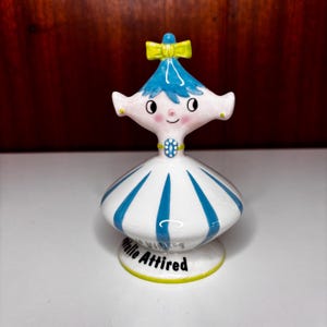 May include: A ceramic figurine of a stylized character with blue hair, a yellow bow, and a white dress featuring vertical blue stripes. The base of the figurine displays the text "Natalie Attired" in black lettering, with a yellow trim along the bottom edge.
