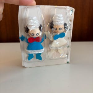 Pode incluir: Duas decorações vintage para árvores de Natal. Uma usa uma roupa azul com uma gravata borboleta vermelha, a outra uma roupa branca com um laço azul. Ambos têm cabelo branco e olhos pretos.