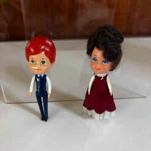 May include: Two vintage miniature dolls. One doll has red hair and is wearing a dark blue suit with a white shirt. The other doll has dark brown hair and is wearing a burgundy dress with a white lace trim.