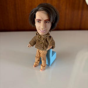 May include: A small doll with a large head, wearing a brown jacket with gold buttons and striped pants. The doll has dark hair and is standing on a small blue platform. The doll's facial features are detailed.