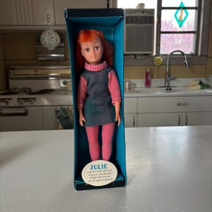 Puede incluir: Una muñeca vintage llamada Julie en una caja azul. La muñeca tiene el pelo rojo, una camisa a rayas azules y blancas, una falda azul y pantalones a rayas rojas y blancas. La caja dice "Julie Twisting Waist Fully Jointed Rooted Hair Sits or Stands".