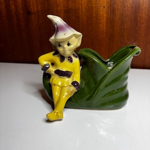 May include: A vintage ceramic planter featuring a whimsical elf figure. The elf is dressed in a yellow outfit with purple accents and a matching hat. The planter is a deep green leaf shape, creating a charming decorative piece.