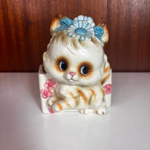 May include: A ceramic cat planter featuring a white and orange tabby kitten with large blue eyes, long eyelashes, and a pink nose. The kitten wears a blue bow with a central flower on its head. The rectangular base is decorated with small pink flowers.