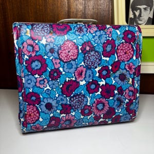 Könnte beinhalten: Vintage Koffer mit floralem Muster. Der Koffer zeigt ein Design aus rosa, blauen und lila Blumen auf hellblauem Hintergrund. Er hat einen weißen Griff und ist rechteckig. Der Koffer ist wahrscheinlich aus einem strapazierfähigen Material.